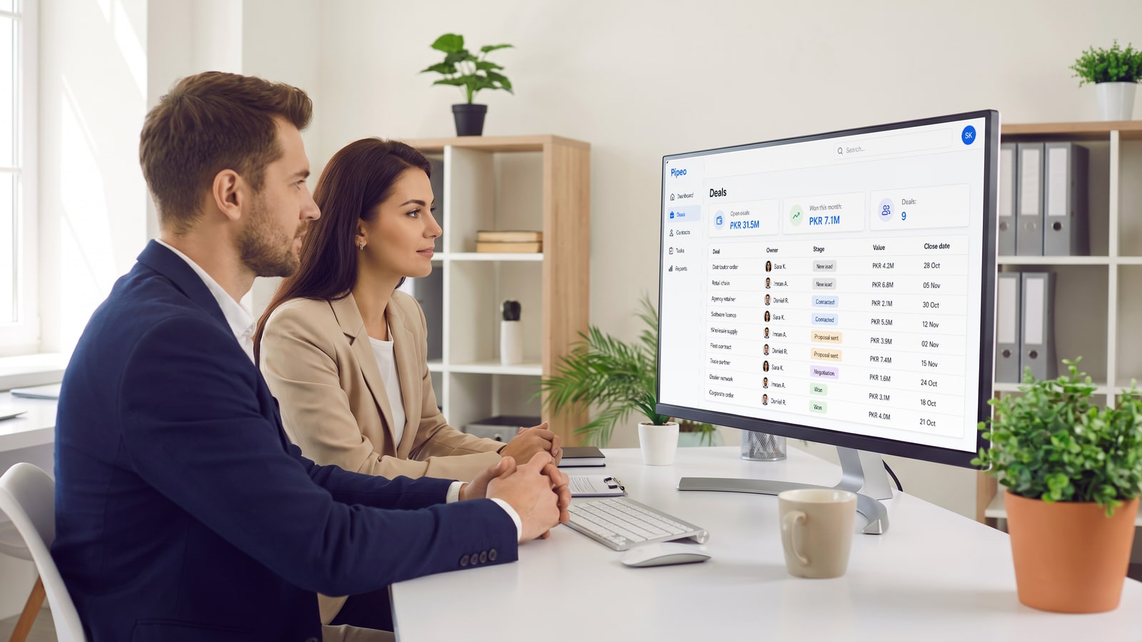 Two colleagues at a shared desk in a bright open-plan office reviewing a sales CRM deals table with stages and values on a large monitor
