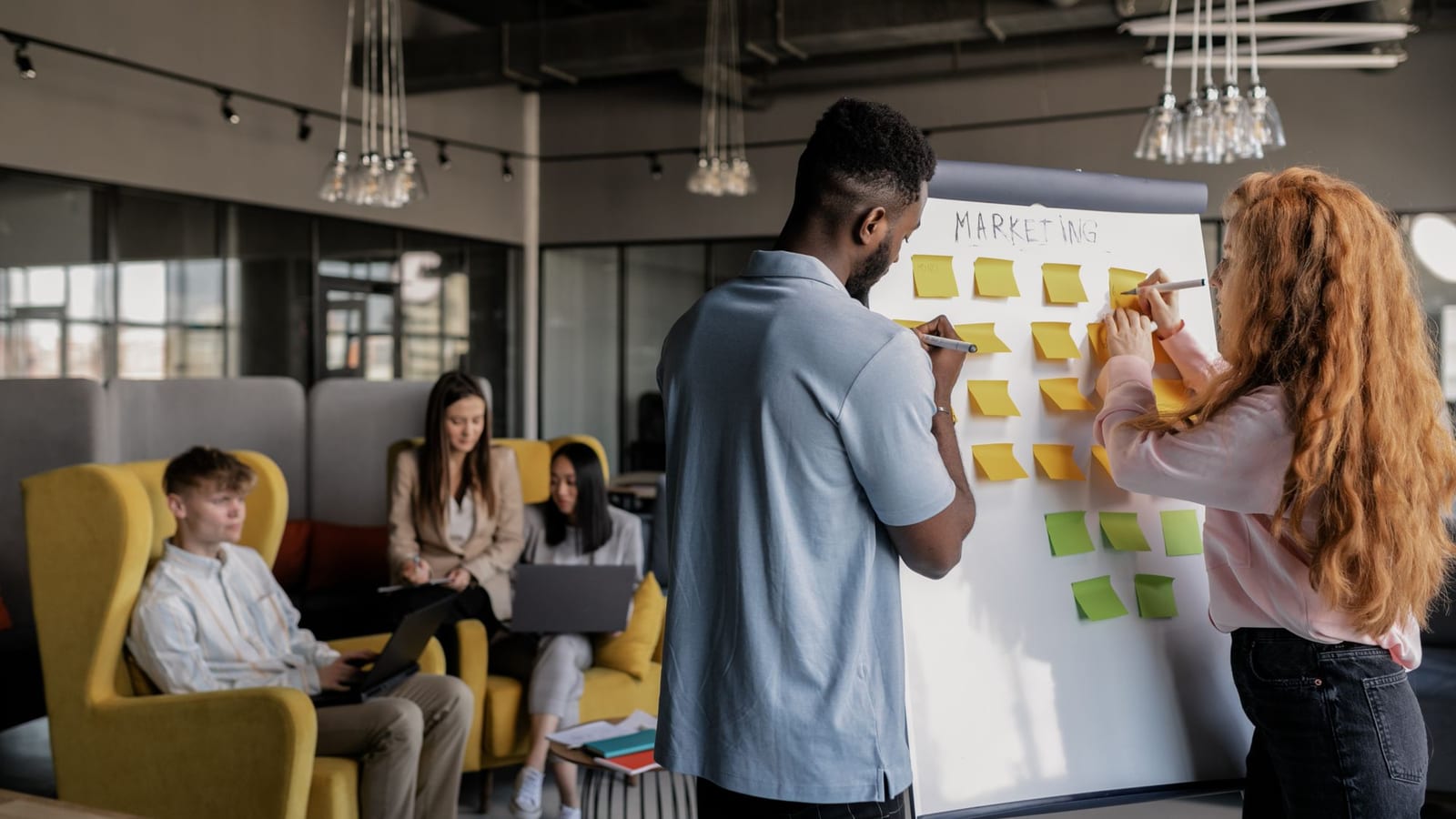 A man and a woman putting sticky notes on a marketing whiteboard while colleagues sit behind them