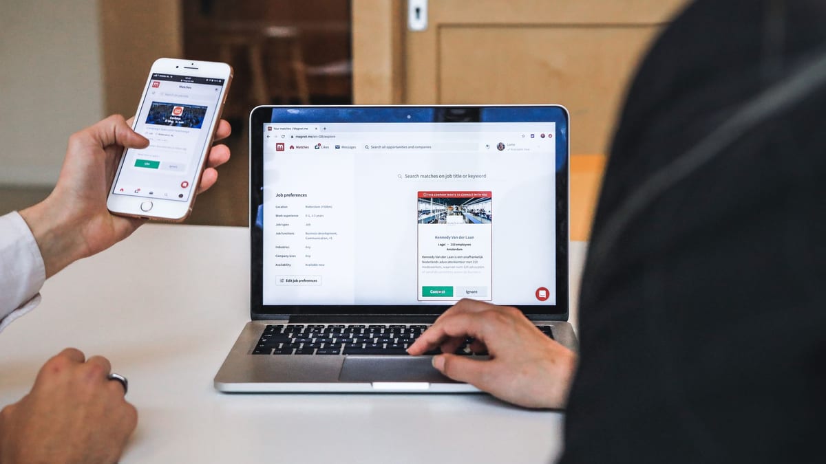 A hand holding a phone beside a laptop showing the same website