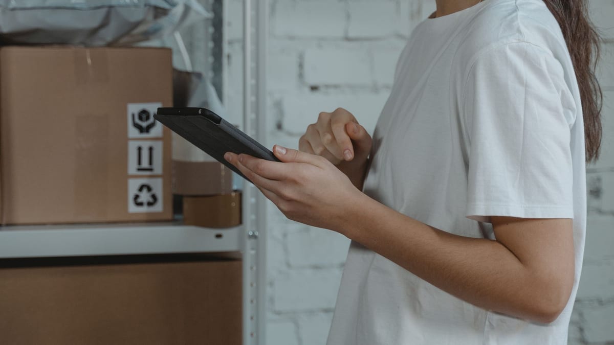A person in a white t-shirt checking stock on a tablet beside shelves of boxes